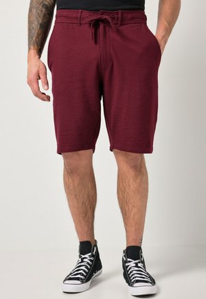 Uomo con pantaloncini rossi scuri strutturati con coulisse e tasche frontali, indossa scarpe da ginnastica nere alte, in piedi davanti a uno sfondo semplice.