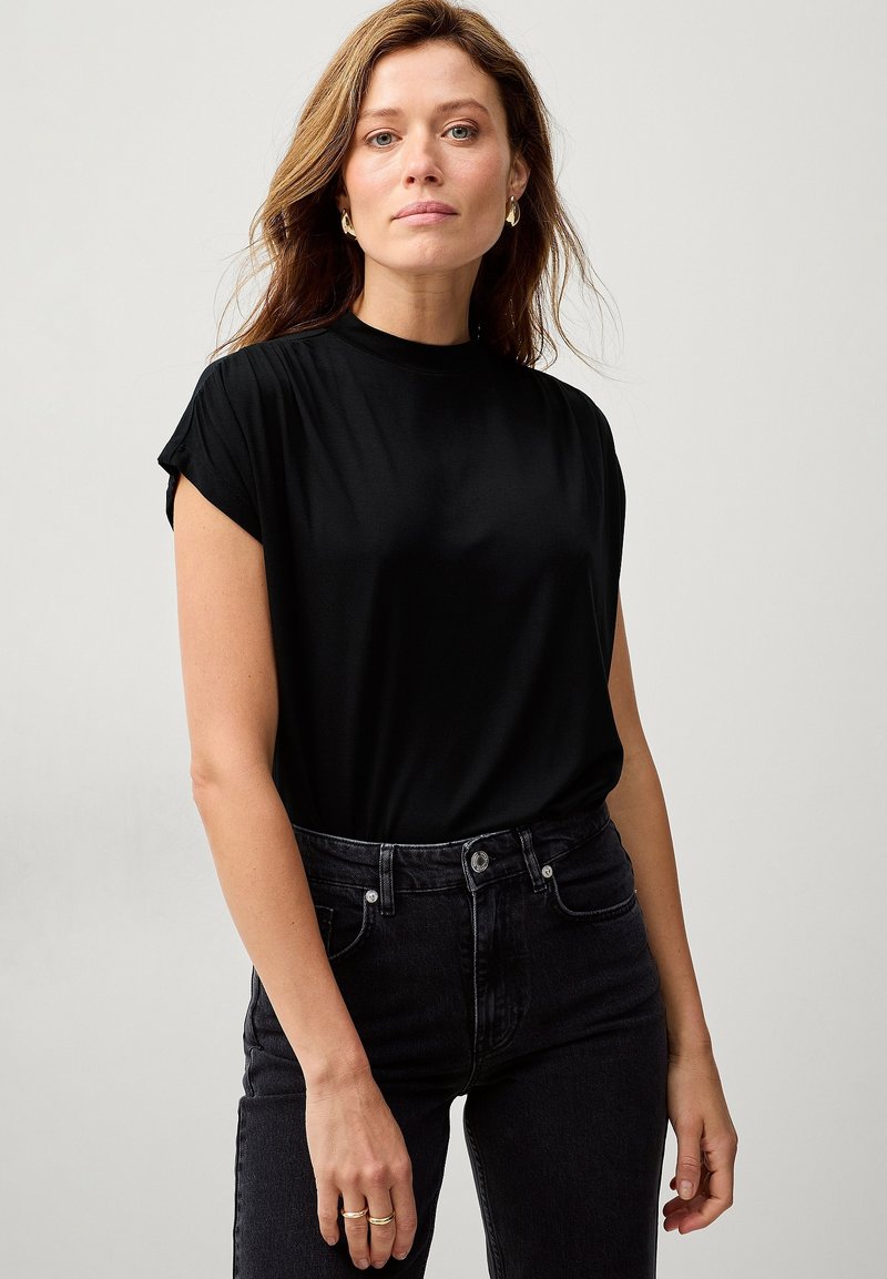 Sort kortærmet skjorte med høj hals, lavet af glat stof. Bæres med mørke denim jeans. Simpelt design uden synlige mønstre.