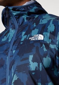 Marinblå jacka med en texturerad, abstrakt mönstring i nyanser av teal. Har en hög krage, justerbar huva och en vit logotyp på bröstet.