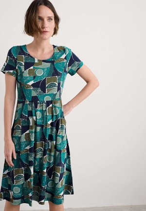 Korte mouwen, jurk tot de knie in teal, marineblauw en olijfgroen. Heeft een geometrisch patroon met cirkels en rechthoeken. Zacht stof met zakken.