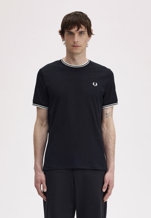 Homme portant un t-shirt noir avec un liseré blanc au niveau du col et des manches, debout de manière neutre devant un arrière-plan clair uni.