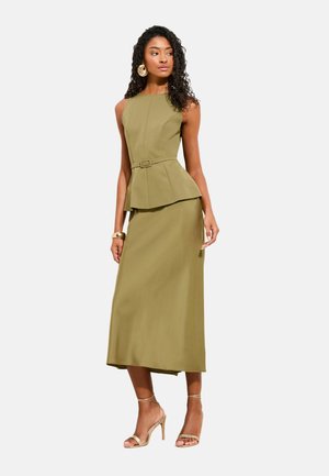 Kvinde med krøllet hår iført olivengrøn ærmeløs top med bælte og peplum samt matchende lang nederdel, guldringeøreringe, armbånd og snørede hæle.