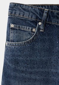 Gros plan de la poche avant d'un jean en denim bleu avec des rivets métalliques et de petits strass bleu foncé dispersés décorant le tissu.