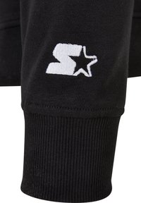 Gros plan sur une manche en tissu noir avec un "S" blanc brodé et un logo étoile au-dessus du poignet côtelé.