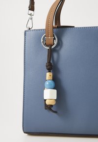 Sac à main bleu texturé avec anse en cuir camel et perles décoratives suspendues en bleu, blanc et beige sur un cordon noir.