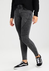 Jeans skinny negros con un lavado desgastado, dotados de paneles acolchados a lo largo de los muslos y cremalleras en los tobillos, combinados con zapatillas negras.