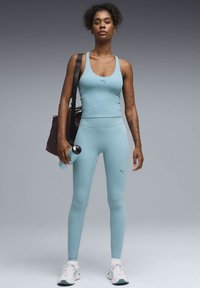 Γυναίκα με αθλητική ενδυμασία ανοιχτού μπλε χρώματος, κρατώντας ένα μπουκάλι νερό και μια μαύρη τσάντα γυμναστηρίου, στέκεται με αυτοπεποίθηση μπροστά σε γκρι φόντο.