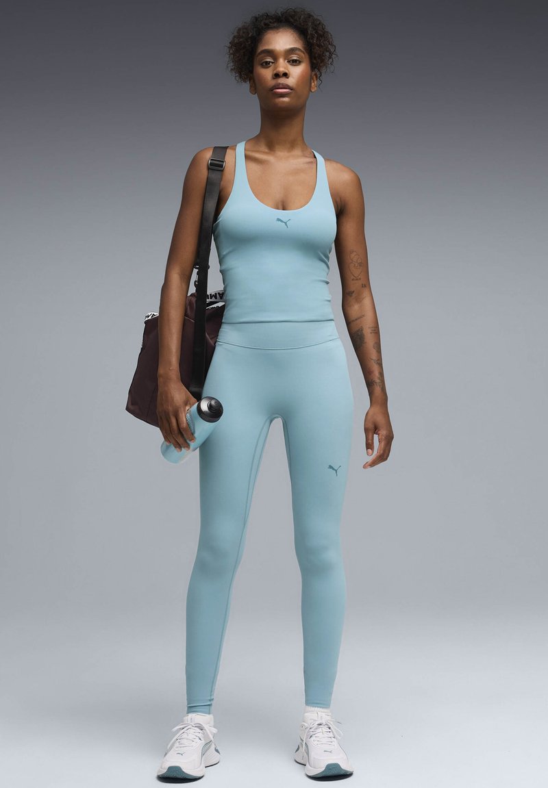 Γυναίκα με αθλητική ενδυμασία ανοιχτού μπλε χρώματος, κρατώντας ένα μπουκάλι νερό και μια μαύρη τσάντα γυμναστηρίου, στέκεται με αυτοπεποίθηση μπροστά σε γκρι φόντο.