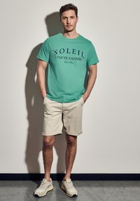 Turkusowa bawełniana koszulka z napisem "SOLEIL" w kolorze granatowym, w połączeniu z beżowymi szortami i beżowymi sneakersami. Prosty krój z podwiniętymi krótkimi rękawami.