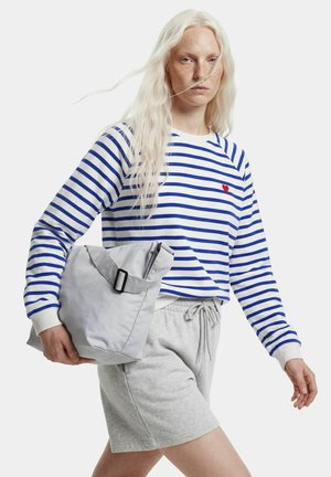 Personne aux longs cheveux blancs portant une chemise rayée bleue et blanche, un short gris, tenant un sac plié gris clair.
