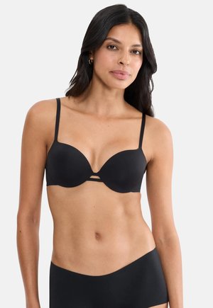 Kvinde med langt mørkt hår iført en sort bh med tynde stropper og matchende sort undertøj mod en ensfarvet baggrund.