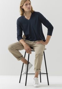 Granatowa koszula z długim rękawem i dekoltem w serek, beżowe spodnie z podwyższonym mankietem i białe trampki, siedząca na drewnianym stołku na jednolitym tle.