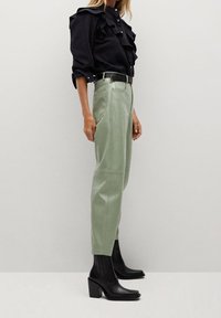 Blusa negra con detalles de volantes, pantalones de cuero verdes ybotines negros de tacón. Los pantalones tienen un corte recto y bolsillos laterales.