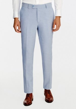 Mężczyzna w jasnoniebieskich spodniach na miarę, białej koszuli i brązowych skórzanych eleganckich butach, stojący na jednolitym białym tle.