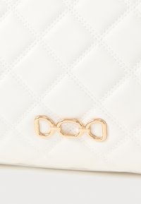 Λευκή τσάντα ώμου με καπιτονέ υφή και λεία, γυαλιστερή επιφάνεια. Η χρυσή μεταλλική λεπτομέρεια στο μπροστινό μέρος έχει σχέδιο με συνδεδεμένους κρίκους.