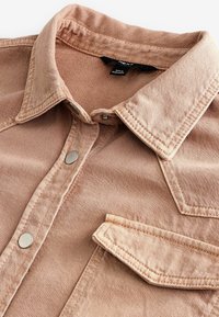 Camicia di jeans rosa chiaro con colletto classico, tasca frontale e bottoni a pressione in metallo. Tessuto liscio con dettagli di cucitura visibili.
