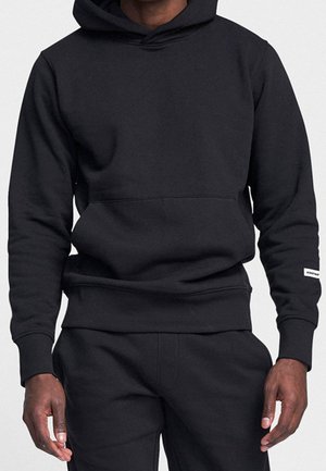 Homme portant un sweat à capuche noir uni et un pantalon noir assorti, debout, bras détendus le long du corps, fond uni.