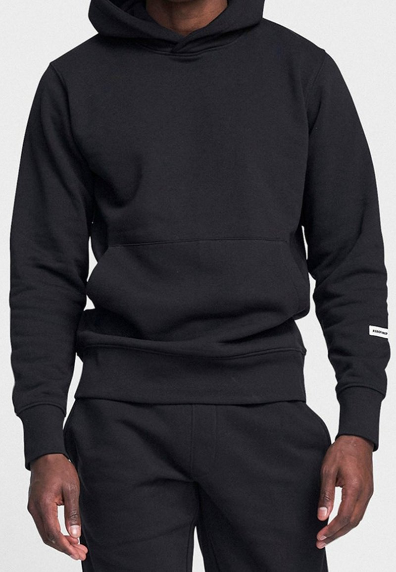 Homme portant un sweat à capuche noir uni et un pantalon noir assorti, debout, bras détendus le long du corps, fond uni.