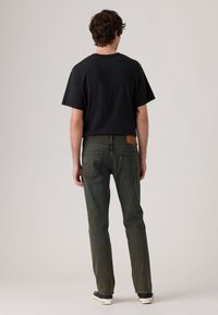 Joven con camiseta negra, jeans oscuros desgastados de color verde y zapatillas negras, de pie de espaldas contra un fondo gris claro liso.