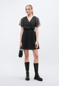 Μαύρο φόρεμα με πουά, V-λαιμόκοψη και κοντά, διαφανή μανίκια· ελαστική μέση· συνδυασμένο με μαύρα μπότες μέχρι το γόνατο και μικρή μαύρη τσάντα.