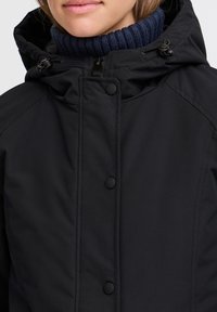 Veste isolante noire avec fermeture éclair, boutons à pression et capuche ajustable. Dotée d'un col côtelé bleu marine pour plus de chaleur.