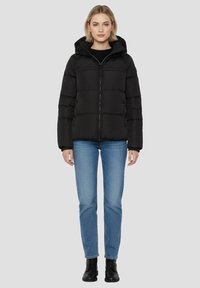 Sort polstret jakke med hætte, med lynlås foran og to sidelommer. Båret over blå jeans og sorte støvler på en neutral baggrund.