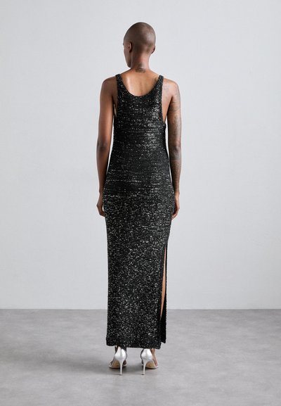 Rochie maxi neagră cu paiete, având o siluetă ajustată, bretele pe umeri, fanta la spate și o textură strălucitoare. Poartă încălțăminte cu toc înalt argintie.