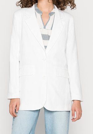 Weißer Leinen-Blazer mit Reverskragen, zwei Knöpfen und seitlichen Taschen, getragen über einem gestreiften Hemd und kombiniert mit hellblauen Jeans.