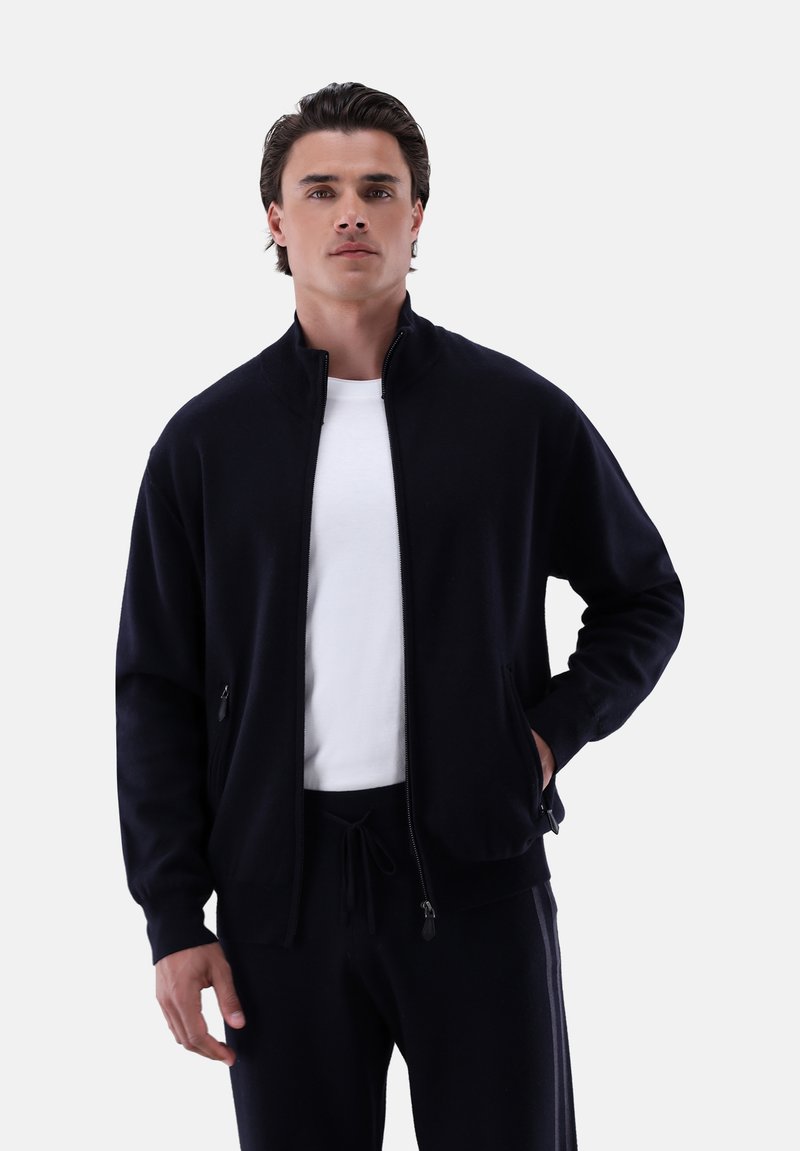 Schwarze Zip-Jacke aus weichem Material mit hohem Kragen, Seitentaschen und gerippten Bündchen. Getragen über einem weißen Hemd und schwarzen Hosen.