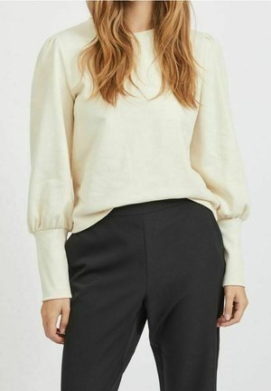 Femme aux longs cheveux châtain clair portant un chemisier crème à manches longues bouffantes et un pantalon noir ajusté.