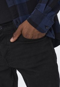 Camicia di flanella a quadri blu e nera con polsini a bottone, indossata sopra pantaloni di jeans neri. La camicia ha una tasca sul petto e una texture morbida.