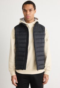 Μαύρο γιλέκο πούφ με οριζόντιο καπιτονέ σχέδιο, μπροστινή φερμουάρ κλεισίματος και λογότυπο από ύφασμα, φορεμένο πάνω από μια κρέμα κουκούλα.