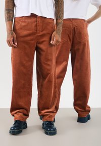 Cordhosen in orange-braun mit vertikalen Streifen, im lässigen Schnitt und mit zwei Gesäßtaschen. Kombiniert mit schwarzen klobigen Schuhen.