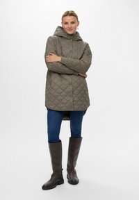 Quiltet, olivengrøn lang jakke med hætte, der har trykknapper, elastiske manchetter og en asymmetrisk kant, parret med blå jeans og brune støvler.