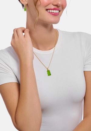 Anhänger in Form eines grünen Gummibärchens an einer Goldkette, getragen zu einem weißen Hemd; verfügt über eine strukturierte Oberfläche und ein verspieltes Design.