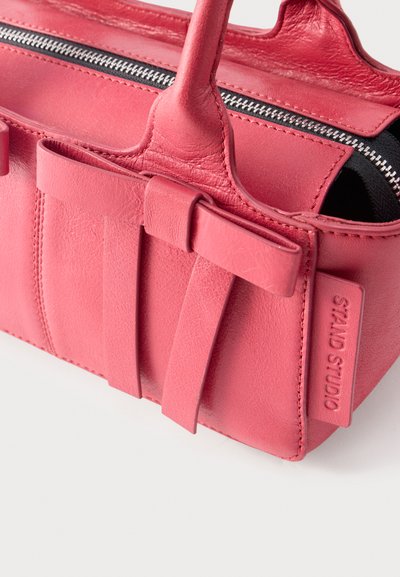 Μικρό ροζ δερμάτινο τσαντάκι χειρός με πάνω λαβές, διακόσμηση φιογκάκι στο μπροστινό μέρος, κύριο διαμέρισμα με φερμουάρ και πλαϊνή ετικέτα με ανάγλυφο το "STAND STUDIO".