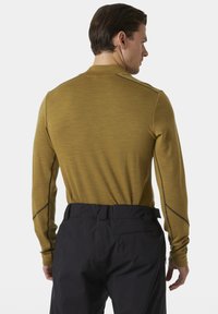 Langsleeve top in olijfgroen, textuurstof met donkere stikselaccenten, gecombineerd met zwarte broek. Focus op de achterkant en pasvorm.
