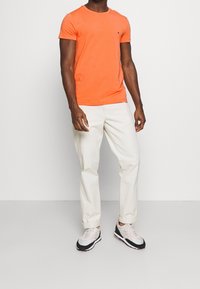 T-shirt arancione a maniche corte, in morbido cotone, con scollatura a girocollo. Indossato con pantaloni beige chiaro e sneakers grigie e bianche da uomo.
