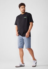 Svart bomull T-skjorte med grønn "Tropica" logo, kombinert med lyseblå denimshorts og hvite sneakers med svarte striper. Ensfarget bakgrunn.