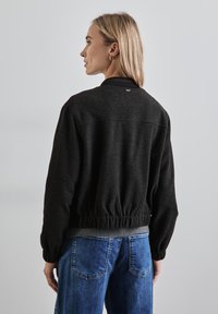Giacca bomber nera in maglia con orlo e polsini a coste, dotata di un discreto logo sul retro. Abbinata a jeans in denim blu.