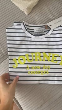 Biely a námornícky pruhovaný tričko s odvážnym žltým textom "JOURNEY" a frázou "Nový deň sa objavuje." Prívesok s názvom značky.