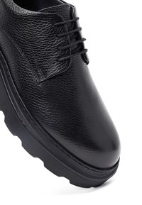 Chaussure en cuir noir avec une finition texturée, dotée d'un bout arrondi, d'un système de laçage et d'une semelle épaisse à nervures.