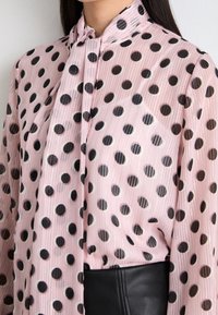 Blouse rose transparente avec de grands pois noirs, col haut et d étail à nouer, présentant une texture côtelée et un léger brillant.