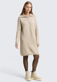 Beżowa dzianinowa sukienka z zamkiem przy kołnierzu, długimi rękawami, prążkową fakturą i bocznymi rozporkami, w zestawieniu z jasnymi kremowymi botkami za kostkę.