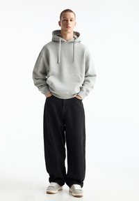 Felpa grigia con cappuccio, tasca a marsupio e stringhe, abbinata a jeans neri larghi e sneakers grigie con suole bianche.