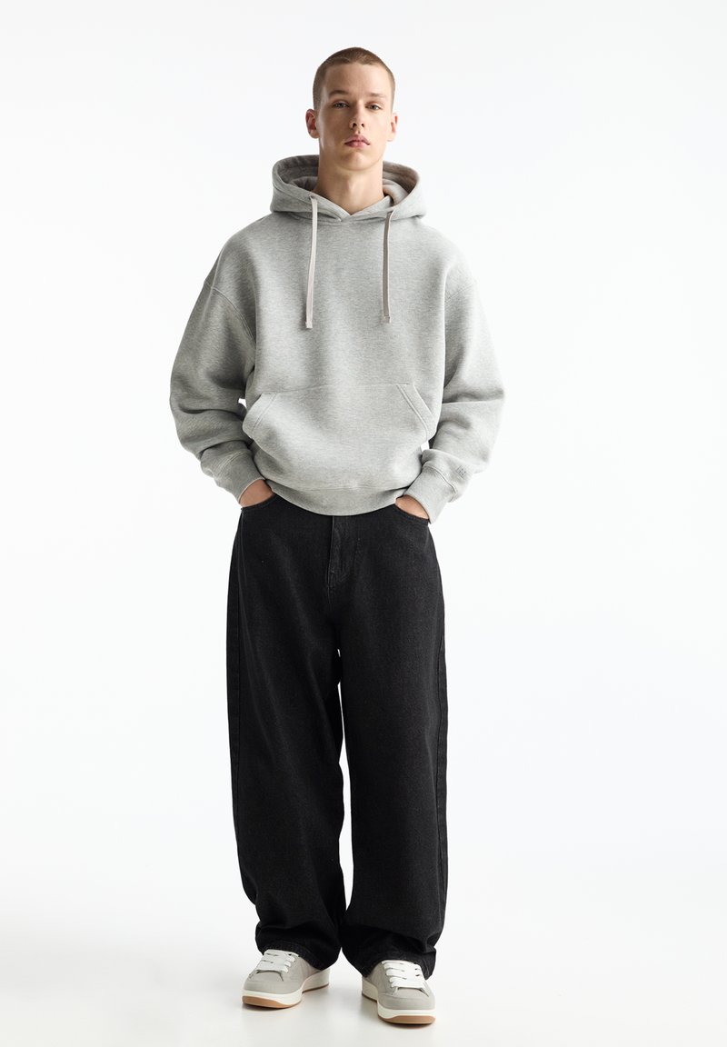 Felpa grigia con cappuccio, tasca a marsupio e stringhe, abbinata a jeans neri larghi e sneakers grigie con suole bianche.