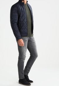 Námořnická prošívaná bunda s stojáčkem, zelený svetr s copánkovým vzorem, šedé slim-fit džíny a černé šněrovací boty.