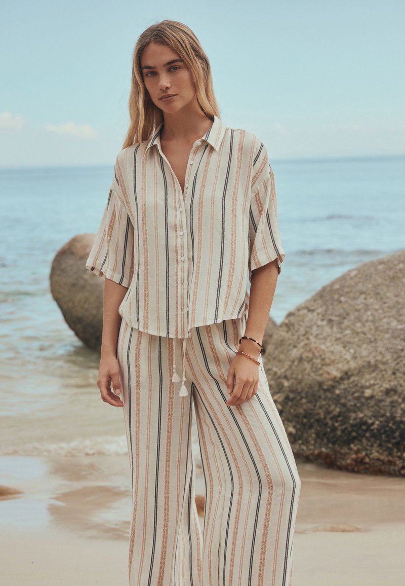 Woman wearing loose, beige striped button-up shirt and matching wide-leg pants standing on a sandy beach near large rocks.