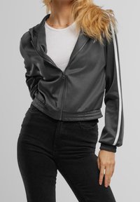 Gray zip-up jacket made of smooth material, featuring white stripes down the sleeves, cropped design, and elasticated hem and cuffs.