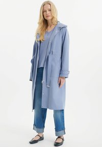 Hellblauer, langer Regenmantel mit Kapuze, Frontreißverschluss, Zugband in der Taille und Druckknöpfen. Wird über einem blauen Oberteil getragen, kombiniert mit hochgekrempelten Jeans.